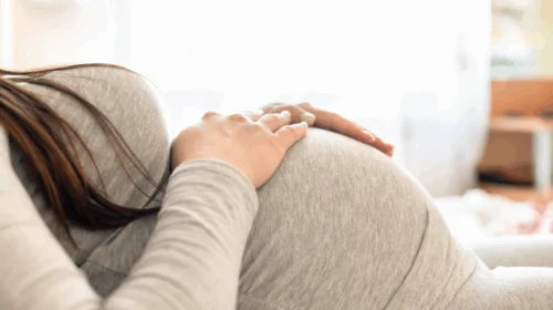 Mulher grávida deitada com as mãos apoiadas na barriga, usando roupa cinza.