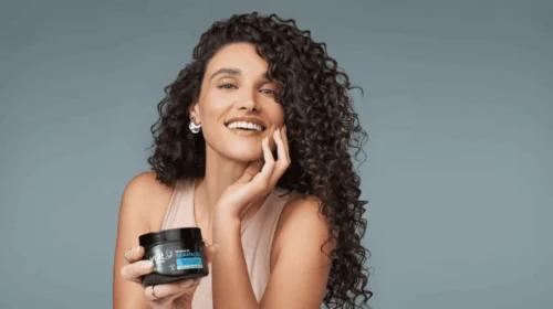 Mulher sorridente com cabelos cacheados segura produto Vult Recarga de Hidratação.