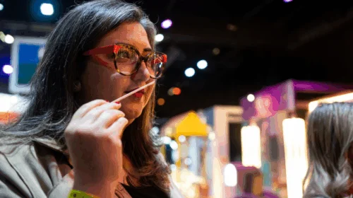 Mulher com óculos e cabelos grisalhos cheira uma fita olfativa em um evento.