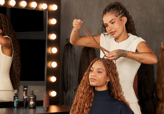 Duas mulheres em um estúdio. Uma trancista com tranças finas e tatuagens faz o cabelo de outra mulher com tranças longas e castanhas.