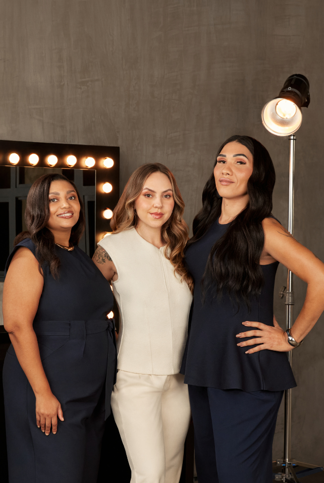 Três mulheres posam em um estúdio, duas vestindo azul-marinho e uma vestindo creme, em frente a um espelho de camarim e um refletor.