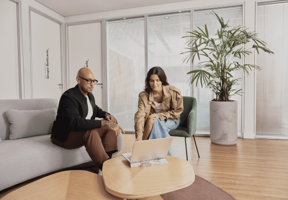 Um homem e uma mulher em uma sala de estar com janelas e plantas. Eles estão sentados em um sofá e uma poltrona, olhando para um laptop.
