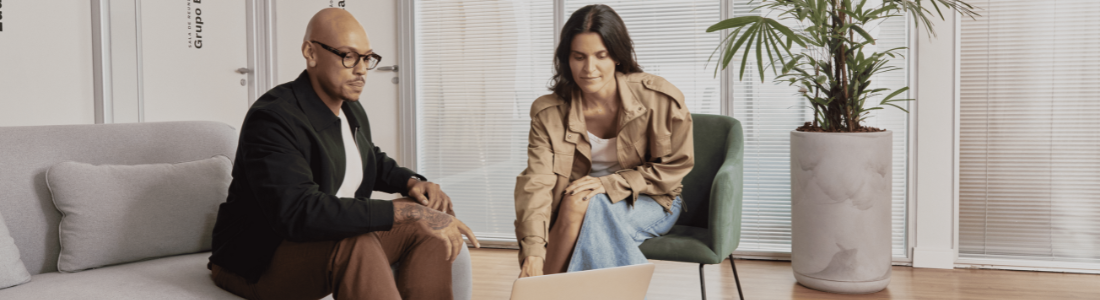 Um homem e uma mulher em uma sala de estar com janelas e plantas. Eles estão sentados em um sofá e uma poltrona, olhando para um laptop.