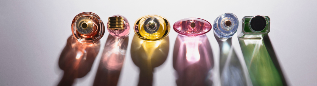 Foto superior das tampas de seis frascos de perfume coloridos (vermelho, rosa, amarelo, pink, azul, verde) em fundo branco com sombras longas.