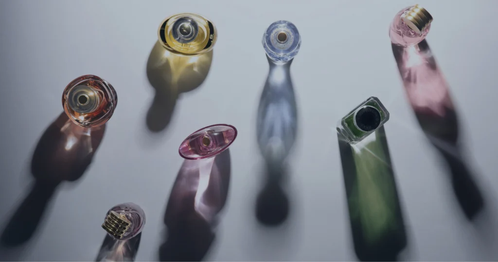 A imagem mostra diversos frascos de vidro em tons multicoloridos, dispostos sobre um plano de fundo neutro na cor cinza.