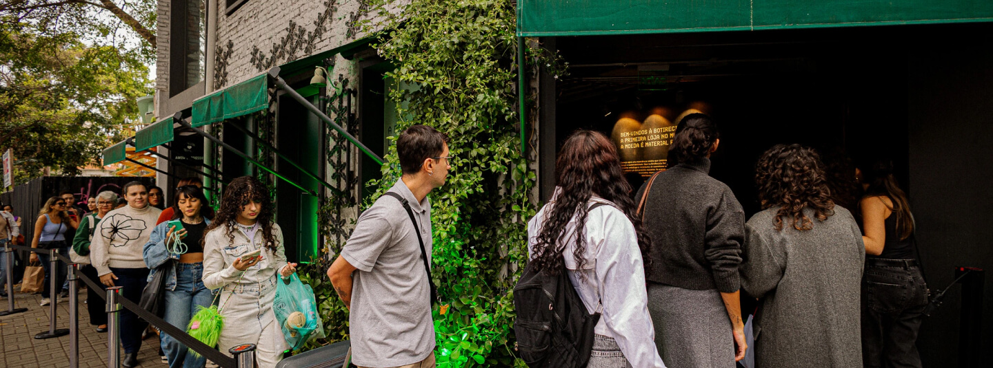 Uma fotografia em plano médio e ângulo aberto captura uma fila de pessoas ao longo da calçada aguardando para entrar em um estabelecimento comercial com fachada escura e detalhes verdes. À direita, um grupo de pessoas é visto de costas, incluindo uma mulher de jaqueta branca e mochila preta, e outras vestidas com casacos cinzas, concentradas diante da entrada escura e sob um toldo verde, onde se projeta um texto iluminado na parede interna. À esquerda, a fila se estende em direção ao fundo da imagem, delimitada por organizadores de fluxo com fitas pretas, mostrando jovens caminhando e conversando, como uma mulher de jaqueta jeans clara olhando para o celular e um homem de camiseta polo cinza e óculos em perfil. A arquitetura do local apresenta paredes de tijolos pintados de cinza, trepadeiras verdes que sobem pela estrutura, e árvores ao fundo que emolduram a movimentação urbana em um dia claro.