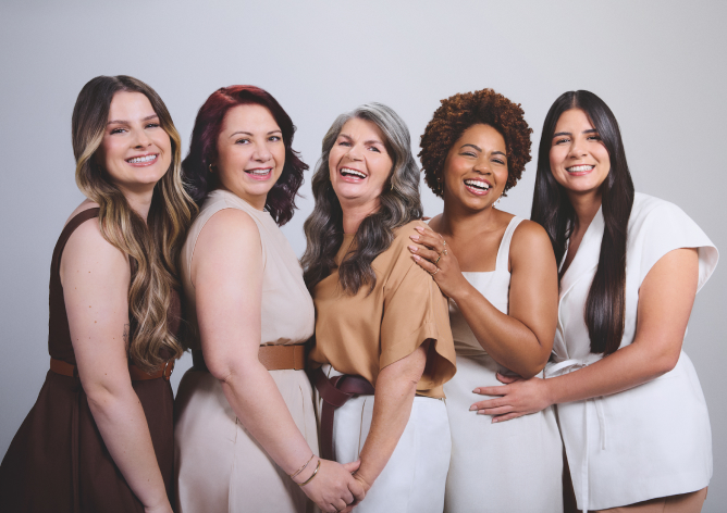 Uma fotografia em plano médio exibe cinco mulheres de diferentes idades, etnias e biotipos alinhadas lado a lado, sorrindo e rindo de forma descontraída em um estúdio com fundo cinza-claro. Da esquerda para a direita, a primeira mulher tem cabelos longos castanhos com mechas loiras e veste um vestido marrom-escuro; a segunda tem pele clara, cabelos avermelhados na altura dos ombros e usa um vestido bege-claro com cinto largo; a terceira, ao centro, é uma mulher mais madura de cabelos longos grisalhos ondulados, que sorri amplamente vestindo uma blusa caramelo e calça branca; a quarta é uma mulher negra de cabelos crespos curtos em tom castanho, que apoia a mão no ombro da colega e veste uma blusa branca de alças; a quinta mulher possui cabelos longos, pretos e lisos, e veste um colete de alfaiataria branco com amarração na cintura. O grupo demonstra forte conexão e cumplicidade por meio de abraços discretos e mãos dadas, exaltando a diversidade e a beleza natural.