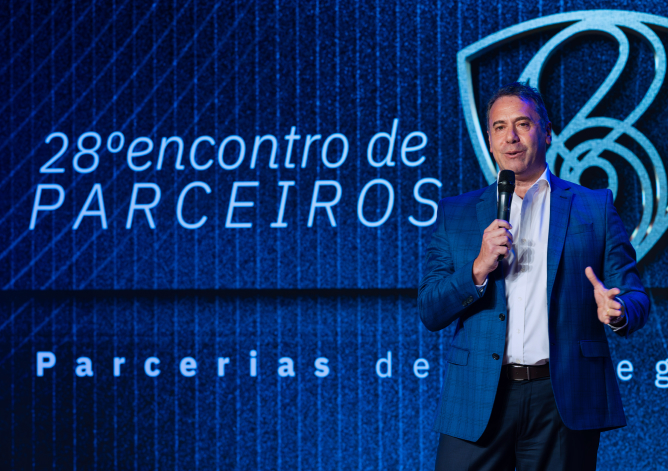 Uma fotografia em plano médio captura um homem de pele clara e cabelos curtos grisalhos discursando no palco de um evento corporativo. Posicionado à direita do enquadramento, ele veste um paletó xadrez em tom azul-escuro sobre uma camisa social branca de gola aberta, calça social escura e cinto de couro marrom. O homem sorri moderadamente enquanto segura um microfone preto com a mão direita e gesticula com a mão esquerda aberta. Ao fundo, um grande painel digital texturizado em azul-escuro exibe à esquerda a inscrição em letras brancas "28º encontro de PARCEIROS" e, na parte inferior, o início da frase "Parcerias de...". No canto superior direito, destaca-se o logotipo estilizado do Grupo Boticário em linhas metálicas prateadas e iluminadas.