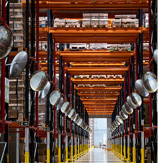 Uma fotografia vertical em perspectiva de ponto central captura um longo e organizado corredor interno de um centro de distribuição ou armazém logístico de grande porte. Em ambos os lados do corredor com piso cinza polido e reflexivo, erguem-se imponentes estruturas metálicas de paletização (porta-paletes) pintadas em tons de laranja vibrante nas vigas horizontais e preto com detalhes vermelhos nas colunas verticais. Os níveis superiores dessas prateleiras estão repletos de paletes de madeira carregados com caixas de papelão uniformes e amarradas.