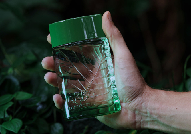 Uma fotografia em plano detalhado mostra uma mão masculina segurando na diagonal um frasco do perfume "Arbo", de O Boticário, contra um fundo escurecido de folhagens verdes naturais. O frasco é retangular, feito de vidro transparente e liso na frente, revelando em relevo interno e na parte posterior o desenho estilizado das nervuras de uma grande folha.