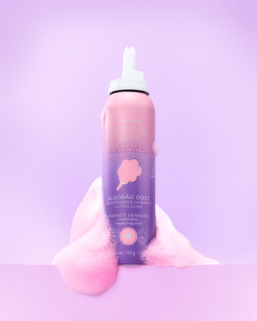 Uma fotografia de estúdio com foco centralizado exibe um frasco de alumínio aerossol do produto "Sabonete em Mousse Corporal" da linha "Cuide-se Bem Doces Delírios", de O Boticário, posicionado verticalmente contra um fundo infinito lilás-claro. O frasco possui um degradê suave que vai do rosa-pastel no topo ao roxo na base, trazendo no centro a ilustração de um algodão-doce rosa no palito, seguida pelas inscrições "Algodão Doce" e "Algodão Doce de Banho". O topo do frasco conta com uma válvula bico injetora de plástico branco, desenhada para criar espuma. Na base do frasco, uma grande quantidade de espuma densa, aerada e de tonalidade rosa-claro transborda para os lados e para a frente sobre uma superfície plana também lilás, simulando a textura macia do produto e reforçando a temática lúdica e doce do cosmético.
