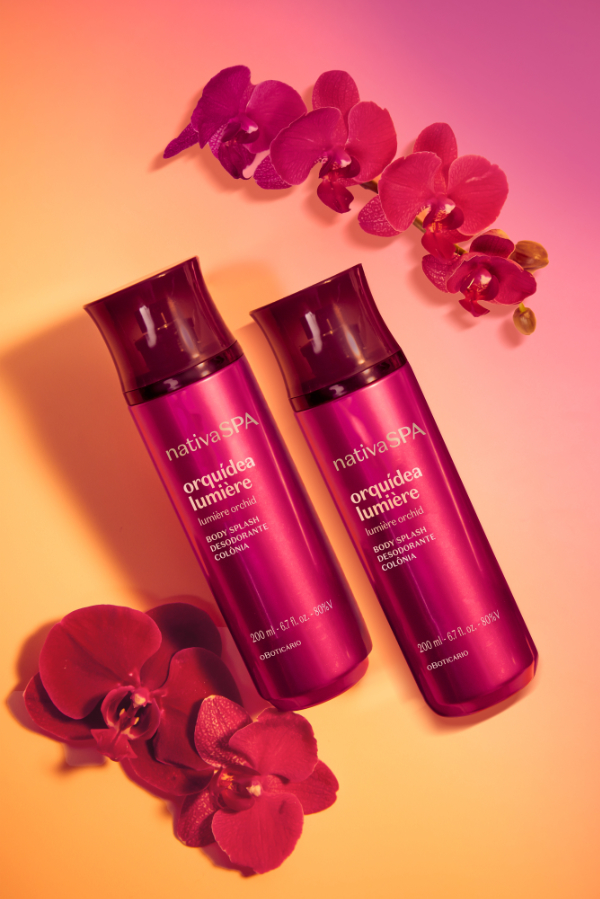 Uma fotografia artística com foco centralizado exibe dois frascos de "Body Splash Desodorante Colônia Orquídea Noire", da linha "Nativa SPA", de O Boticário, dispostos diagonalmente lado a lado sobre uma superfície lisa. Os frascos são cilíndricos e alongados, feitos de material translúcido em um tom magenta ou roxo-vibrante com acabamento brilhante, finalizados por tampas plásticas em formato côncavo de cor roxo-escura. O rótulo frontal traz as inscrições da marca e do produto em letras brancas e prateadas, indicando o volume de 200 ml. Decorando a composição, flores de orquídea em tons de roxo e vinho estão posicionadas ao redor dos produtos: uma ramificação arqueada adorna a parte superior, enquanto duas flores soltas aparecem no canto inferior esquerdo. O fundo apresenta um fundo em degradê suave que transita do amarelo-alaranjado quente no canto inferior esquerdo para um rosa-vibrante no canto superior direito, criando uma iluminação tropical e sofisticada.