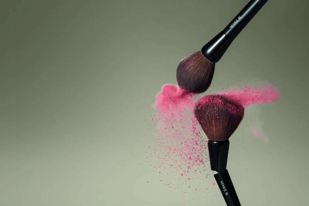 A imagem mostra dois pincéis da linha MakeB depositando uma névoa de blush rosa sobre um plano de fundo neutro na cor marrom.