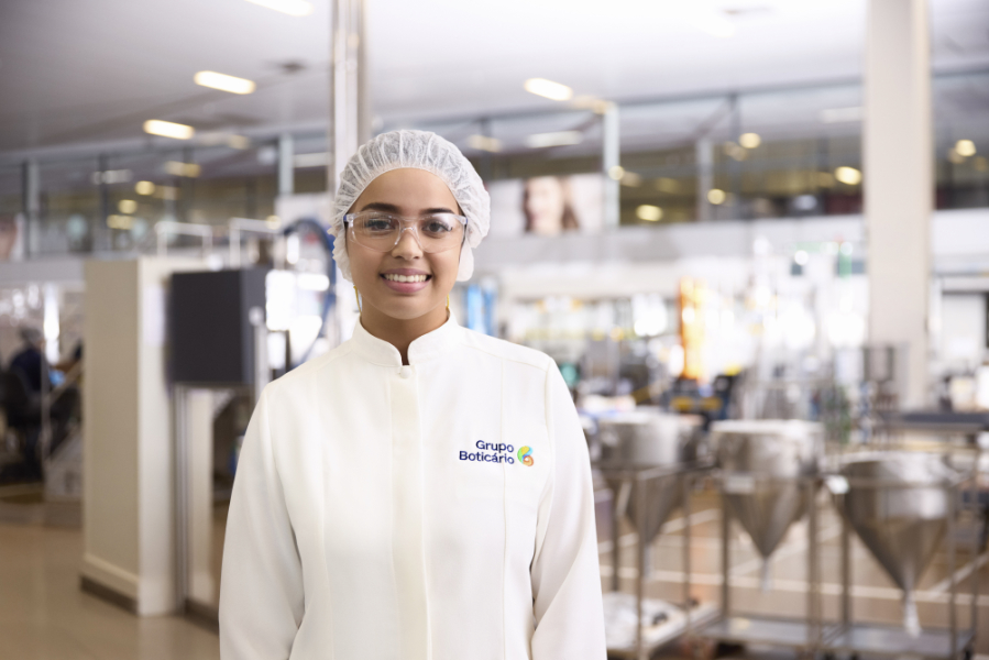 Uma fotografia em plano médio e ângulo frontal destaca uma mulher jovem de pele morena clara, sorrindo gentilmente em direção à câmera no interior de uma unidade industrial. Ela veste um jaleco branco de gola padre com o logotipo do "Grupo Boticário" bordado em azul, amarelo e verde no lado esquerdo do peito, além de usar óculos de proteção transparentes e uma touca descartável branca cobrindo os cabelos. O ambiente fabril ao fundo está ligeiramente desfocado, revelando uma estrutura ampla e iluminada com pisos claros, colunas e maquinários em aço inoxidável, como grandes tanques cônicos de mistura posicionados à direita, reforçando o cenário de um laboratório ou linha de produção de cosméticos.