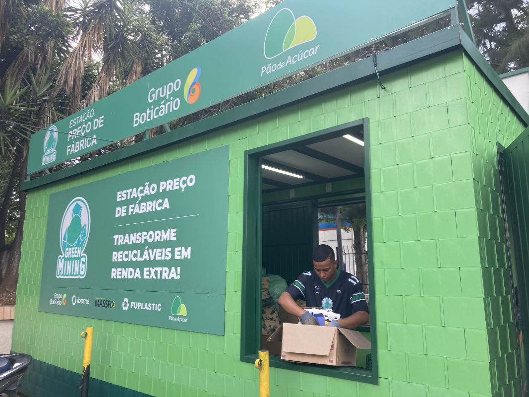 Uma fotografia em plano médio e ângulo levemente contra-plongée mostra um quiosque de reciclagem sustentável pintado em tons de verde, integrado a um cenário externo com árvores ao fundo. O quiosque possui paredes externas revestidas com placas modulares verde-alface e um grande painel frontal em verde-escuro, onde se lê a inscrição "ESTAÇÃO PREÇO DE FÁBRICA - TRANSFORME RECICLÁVEIS EM RENDA EXTRA!", acompanhada pelo logotipo da "Green Mining" e marcas parceiras na base, como Grupo Boticário e Pão de Açúcar. 