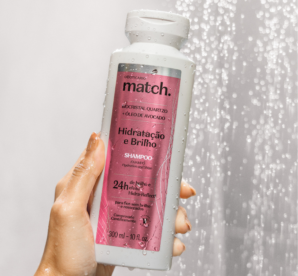 Uma fotografia em plano detalhado e vertical mostra uma mão segurando um frasco branco do shampoo "Match Hidratação e Brilho", de O Boticário, posicionado na diagonal em frente a uma ducha de água corrente. O frasco é retangular e opaco, com cantos arredondados, e traz um rótulo frontal retangular em tom rosa-quartzo metalizado, onde se lê a marca "match." em letras minúsculas pretas, seguida pelas descrições "BIOCRISTAL QUARTZO + ÓLEO DE AVOCADO", "SHAMPOO" e o destaque "24h de brilho e efeito". Tanto a mão quanto o frasco estão salpicados por pequenas gotas d'água. À direita, um fluxo intenso e brilhante de água em movimento cai verticalmente com um efeito desfocado (bokeh), criando pequenas esferas luminosas que reforçam a atmosfera refrescante de um banho.
