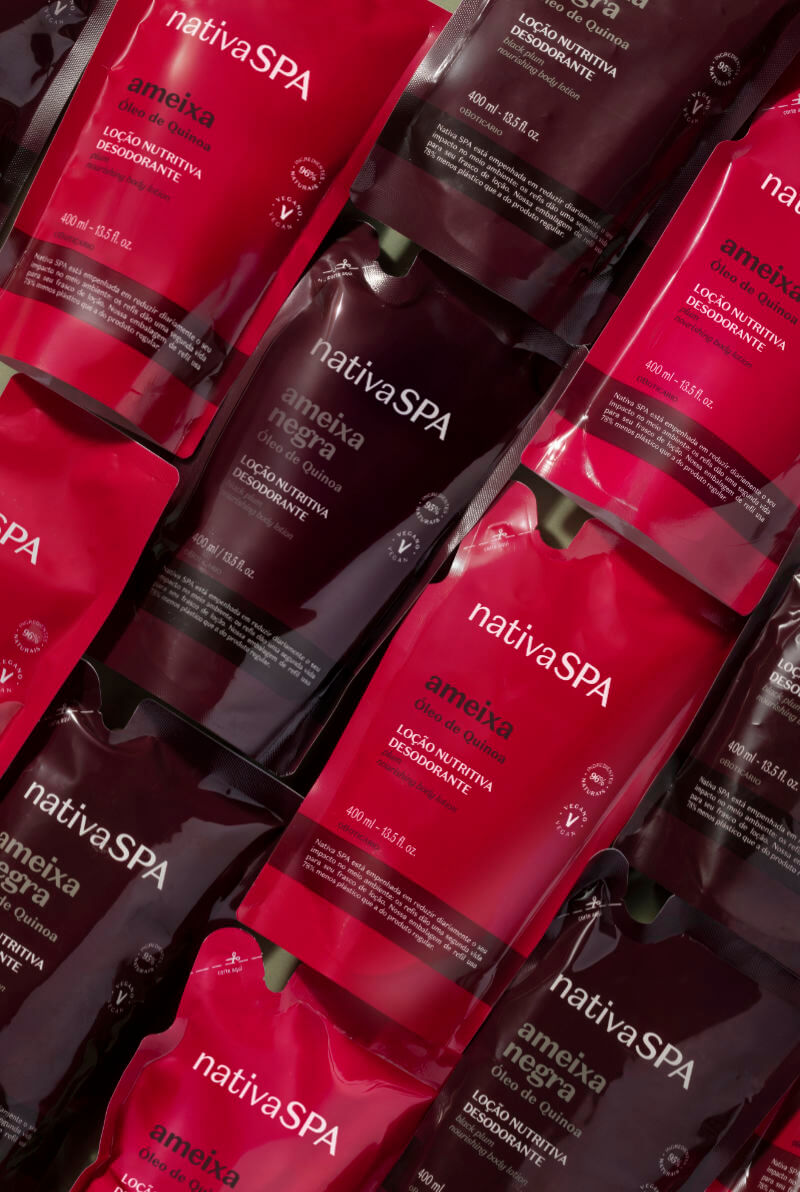Uma fotografia em plano detalhado e com ângulo ligeiramente diagonal exibe diversas embalagens de refil em formato pouch da linha "Nativa SPA", de O Boticário, dispostas de maneira sobreposta. As embalagens dividem-se em duas variações de cores e fragrâncias: algumas são de um tom vermelho-vivo brilhante, correspondentes à fragrância "ameixa", e outras possuem um tom roxo-escuro profundo ou bordô, da fragrância "ameixa negra".