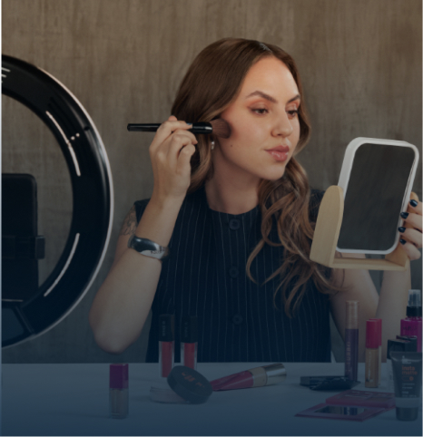 Uma fotografia em plano médio mostra uma mulher de cabelos castanhos claros e ondulados sentada em frente a uma mesa branca, aplicando maquiagem. Sobre a mesa, há uma grande variedade de produtos cosméticos espalhados, como gloss labial, bases, corretivos, paletas de sombras e um copo de vidro transparente com vários pincéis de maquiagem.