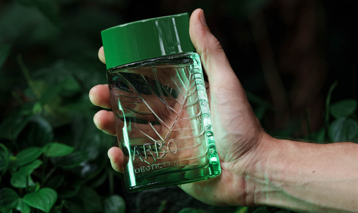 Uma fotografia em plano detalhado mostra uma mão masculina segurando na diagonal um frasco do perfume "Arbo", de O Boticário, contra um fundo escurecido de folhagens verdes naturais. O frasco é retangular, feito de vidro transparente e liso na frente, revelando em relevo interno e na parte posterior o desenho estilizado das nervuras de uma grande folha.
