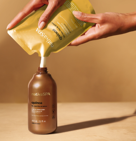 Uma fotografia em plano detalhado e com foco centralizado registra o momento em que duas mãos com unhas bem cuidadas seguram um sachê de refil flexível dourado para reabastecer um frasco de loção hidratante corporal. O frasco cilíndrico de base plana é feito de plástico marrom-acobreado metálico da linha "Nativa SPA Quinoa", de O Boticário, e está posicionado verticalmente sobre uma superfície fosca bege-clara. O bico do sachê dourado, que está virado de ponta-cabeça na parte superior, está perfeitamente acoplado ao gargalo aberto do frasco, transferindo o creme de loção firmadora desodorante. O rótulo frontal do frasco exibe as inscrições em letras pretas e douradas, indicando o volume de "400 ml". O fundo e a iluminação quente em tons terrosos criam uma atmosfera aconchegante, destacando o ato sustentável e consciente do uso de refis na rotina de beleza.