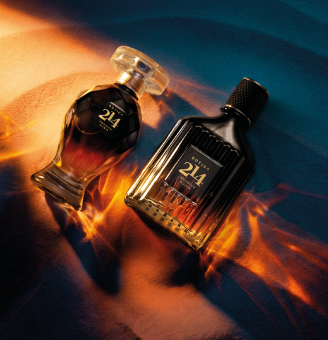Uma fotografia artística em plano zenital (visto de cima) destaca dois frascos de perfume da linha "Botica 214 Arabian Oasis", de O Boticário, deitados diagonalmente sobre uma superfície texturizada que simula dunas de areia fina. À esquerda, encontra-se o frasco feminino, com silhueta curvilínea e lapidada em formato de ânfora, finalizado por uma tampa redonda transparente e achatada. À direita, está o frasco masculino, de formato retangular e imponente com ranhuras verticais e uma tampa cilíndrica preta texturizada. Ambos os frascos são feitos de vidro escuro translúcido com gradiente âmbar na base e exibem a inscrição "BOTICA 214" em letras douradas marcantes. A iluminação dramática e contrastante divide a cena entre um fundo azul-petróleo frio e feixes de luz quente e alaranjada que atravessam os frascos, projetando reflexos luminosos e sinuosos na areia que remetem ao calor do deserto.