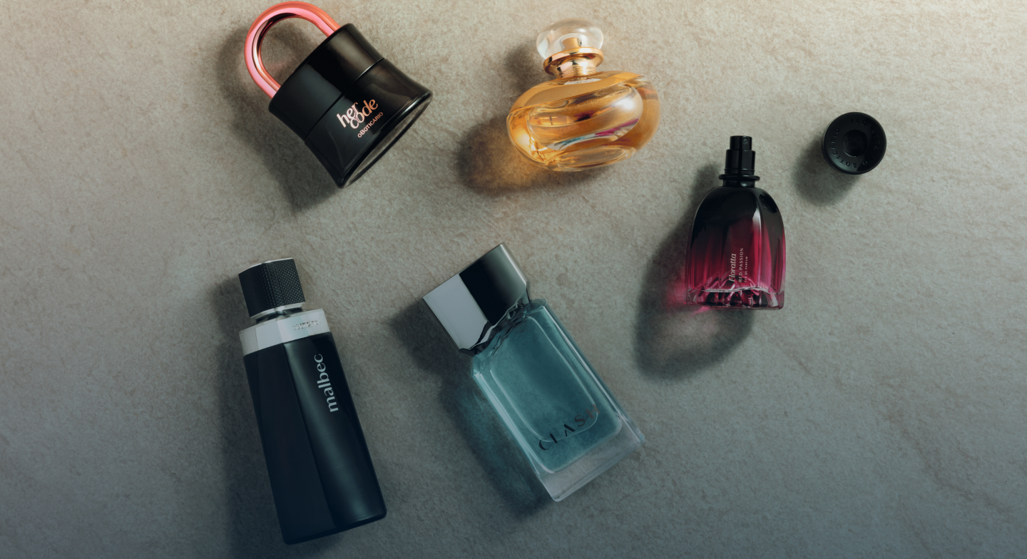 Uma fotografia tirada de um ângulo superior plano (visto de cima) exibe uma coleção de cinco frascos de perfume de diferentes formatos e cores, dispostos sobre uma superfície texturizada bege que imita pedra ou cimento claro. Na foto, há frascos dos perfumes Her Code, Floratta, Malbec e Clash.