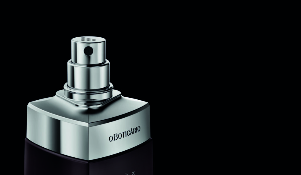 Uma fotografia em plano detalhado e com enquadramento descentralizado destaca a parte superior de um frasco de perfume masculino, posicionado no canto inferior esquerdo contra um fundo liso e minimalista em tom cinza-escuro. O frasco possui ombros robustos de metal prateado polido e espelhado com acabamento geométrico, onde se nota a marca "O Boticário" gravada discretamente em letras maiúsculas pretas na lateral.