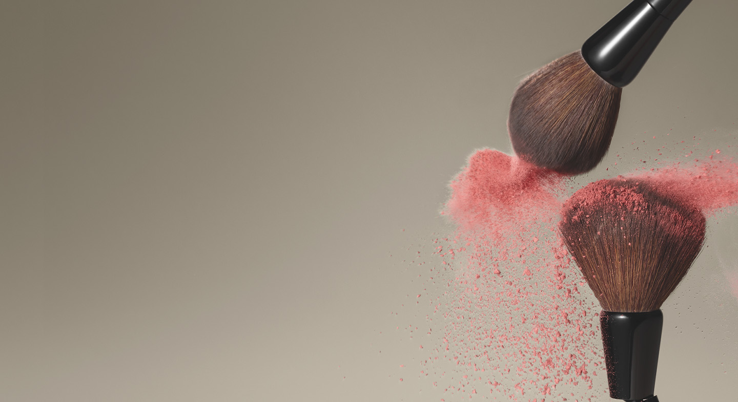 A imagem mostra dois pincéis da linha MakeB depositando uma névoa de blush rosa sobre um plano de fundo neutro na cor marrom.