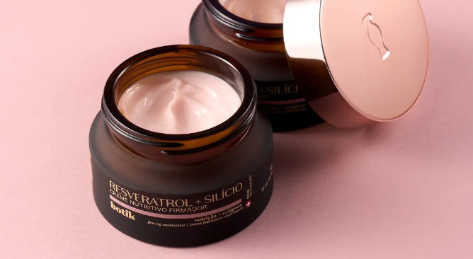 Uma fotografia em ângulo superior exibe dois potes circulares de creme facial da linha "Botik" dispostos sobre uma superfície lisa de cor rosa-antigo. Ambos os potes revelam um creme consistente de tom rosa-claro com leves ondulações na textura; sua embalagem é de vidro fosco escuro com rótulo preto onde se lê, em letras douradas e brancas, "Resveratrol + Silício Creme Nutritivo Firmador", seguido pela marca "botik" e detalhes sobre colágeno e nutrição.