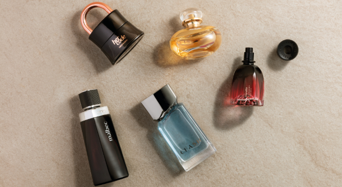 Uma fotografia tirada de um ângulo superior plano (visto de cima) exibe uma coleção de cinco frascos de perfume de diferentes formatos e cores, dispostos sobre uma superfície texturizada bege que imita pedra ou cimento claro. Na foto, há frascos dos perfumes Her Code, Floratta, Malbec e Clash.