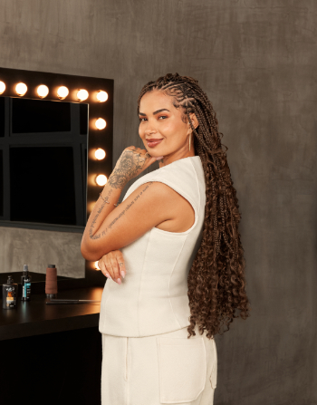 Uma fotografia vertical em plano médio mostra uma mulher de pele morena posicionada de perfil para a esquerda, virando o rosto e sorrindo com confiança em direção à câmera. Ela tem longas tranças nagô e box braids castanhas que se estendem até abaixo da cintura, terminando em pontas cacheadas e volumosas. A mulher veste um conjunto de colete sem mangas e calça na cor off-white, possui tatuagens detalhadas no braço e na mão esquerda, e apoia suavemente o queixo sobre os dedos.