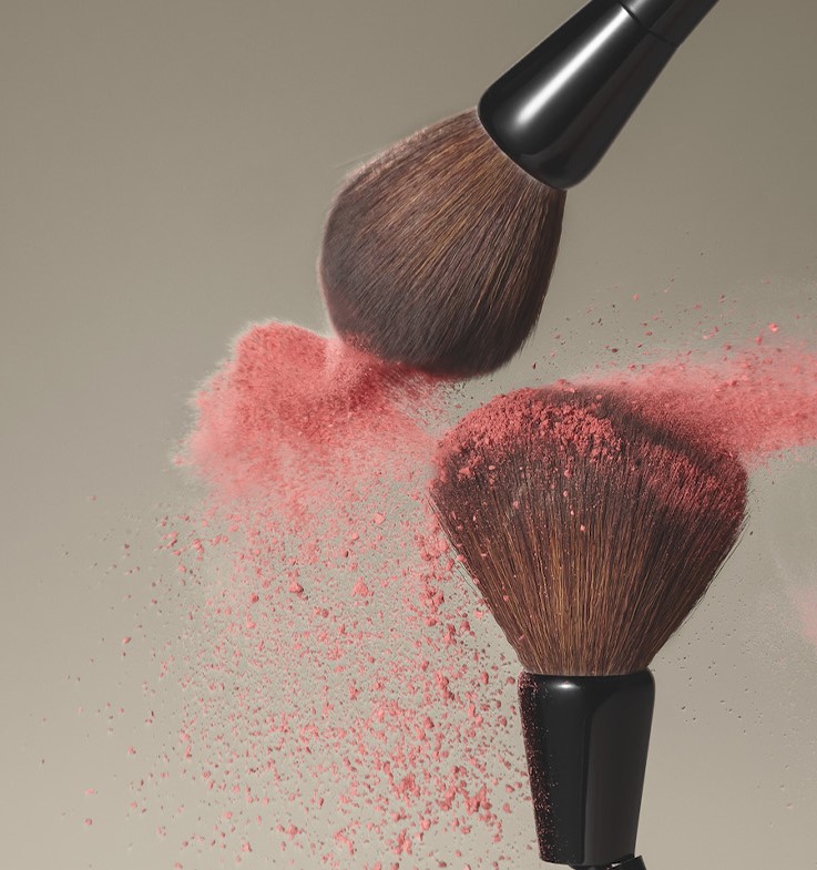 A imagem mostra dois pincéis da linha MakeB depositando uma névoa de blush rosa sobre um plano de fundo neutro na cor marrom.