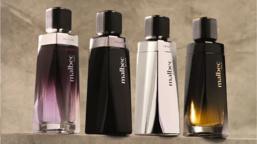 Quatro frascos de perfume Malbec lado a lado sobre uma superfície de pedra cinza. Da esquerda para a direita: Malbec Icon (roxo degrau), Malbec Black (preto fosco), Malbec Magnetic (prateado espelhado) e Malbec Gold (preto com fundo dourado). O fundo é uma parede de pedra cinza neutra.