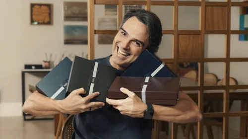 Homem sorridente e de terno segurando quatro caixas de presente.