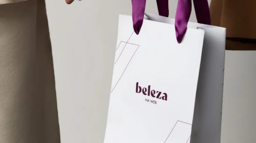 Close-up em ângulo de uma sacola de compras de papel branco da marca 'beleza NA WEB', com alças de fita de cetim roxas. A sacola tem linhas geométricas finas e a marca está centralizada em roxo escuro. As mãos de uma pessoa com roupas em tons de bege seguram a sacola contra um fundo cinza neutro.