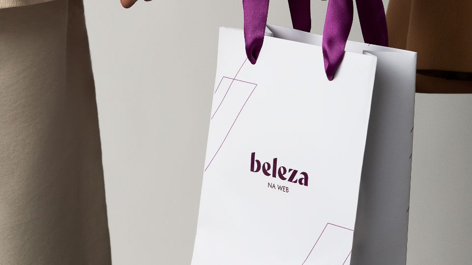 Close-up em ângulo de uma sacola de compras de papel branco da marca 'beleza NA WEB', com alças de fita de cetim roxas. A sacola tem linhas geométricas finas e a marca está centralizada em roxo escuro. As mãos de uma pessoa com roupas em tons de bege seguram a sacola contra um fundo cinza neutro.