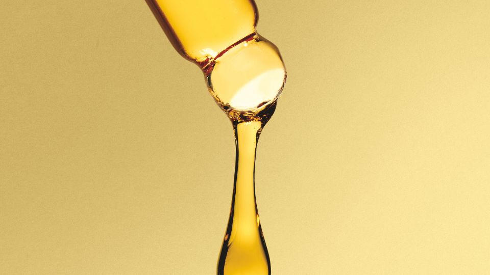 Close-up em macro de um conta-gotas de vidro liberando uma gota espessa e âmbar de óleo cosmético ou sérum contra um fundo dourado fosco.